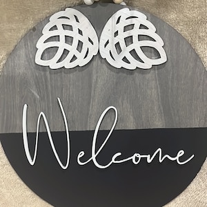 May include: A round wooden welcome sign with a black and gray wood grain finish. The sign has two white, decorative, cutout leaves at the top and the word "Welcome" written in white cursive on the black bottom half.