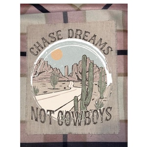 May include: A graphic design featuring a desert landscape with cacti, mountains, and a road. The text "CHASE DREAMS" arches above the image, and "NOT COWBOYS" is below. The design is on a square patch with a frayed edge.