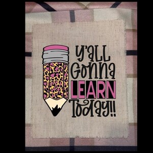 May include: A tan fabric patch with a pencil graphic and the text "Y'all Gonna Learn Today!!". The pencil has a pink eraser, a gray band, and a leopard print design. The text is in black and pink.