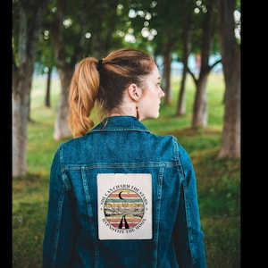 May include: A denim jacket with a patch featuring a desert landscape, a crescent moon, and the text "She Can Charm the Stars, Hypnotize the Moon." The jacket is a medium blue color.
