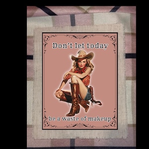 May include: A vintage-style print featuring a cowgirl in a hat and boots, with the text "Don't let today be a waste of makeup." The image has a pink background and a decorative black border, evoking a retro aesthetic.