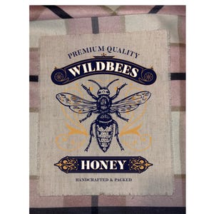 May include: A beige rectangular label with the words "PREMIUM QUALITY WILDBEES HONEY HANDCRAFTED & PACKED" and a detailed illustration of a bee. The design is in navy blue and gold, set against a textured background.
