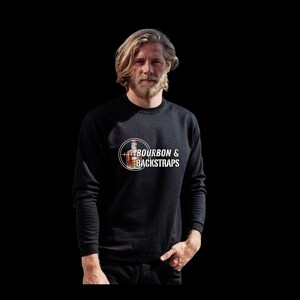 Puede incluir: Sudadera negra con el texto "BOURBON & BACKSTRAPS" en un diseño circular blanco. El diseño incluye una ilustración de una botella de whisky. La sudadera es de manga larga y tiene cuello redondo.