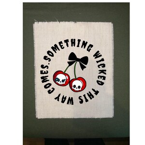 May include: A white fabric patch with a distressed edge features a circular design with the text "SOMETHING WICKED THIS WAY COMES." Two red cherries with skull faces and a black bow are in the center. The patch is on a green background.