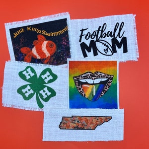 May include: Five fabric patches with different designs. The top left patch is black with a white fish and the text "Just Keep Swimming". The top right patch is white with black text that says "Football Mom" and a football with a heart. The bottom left patch is white with a green four-leaf clover with the letter "H" on each leaf. The bottom right patch is white with a brown outline of the state of Tennessee with the word "Home" inside. The center patch is a rainbow with a leopard tongue sticking out.