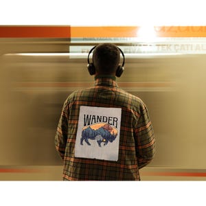May include: A person wearing a green and brown plaid shirt with a patch that says "WANDER" and features a bison illustration. The person is wearing headphones. The background is blurred with orange and brown tones.