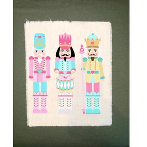 May include: Three nutcracker figurines with pink, blue, and yellow accents. The nutcrackers are wearing hats and have facial features. The nutcracker on the left is holding a drum, the middle nutcracker is holding a drum, and the nutcracker on the right is holding a staff.