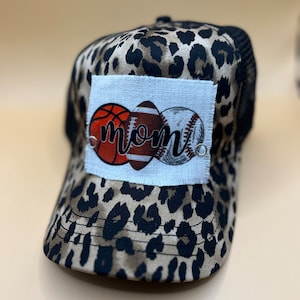 May include: A brown and black leopard print trucker hat with a white patch featuring a basketball, football, and baseball with the word "mom" in black lettering.