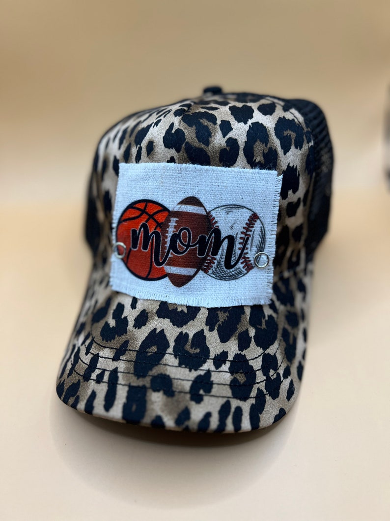 May include: A brown and black leopard print trucker hat with a white patch featuring a basketball, football, and baseball with the word "mom" in black lettering.