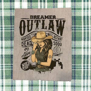 May include: A light gray poster with a Wild West theme. The poster features a woman wearing a cowboy hat, with the words "Dreamer Outlaw" above her. The text "Wanted Dead or Alive" is also present, along with a drawing of a gun.