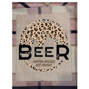 May include: A graphic design featuring the word "BEER" in bold black letters, with the phrase "NEVER BROKE MY HEART" underneath. The design includes a leopard print arch and is set against a neutral background.