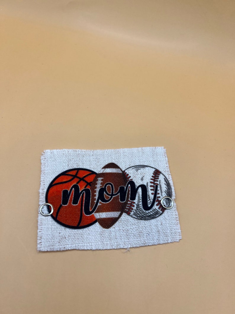 May include: A white fabric patch with a basketball, football, and baseball design. The word "MOM" is written in black cursive across the balls.