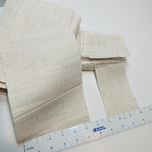 May include: A collection of beige fabric squares and strips, some neatly stacked, others with frayed edges. A ruler with inch markings is placed alongside the fabric, showing measurements up to 11 inches. The fabric appears to be linen or a similar natural fiber.