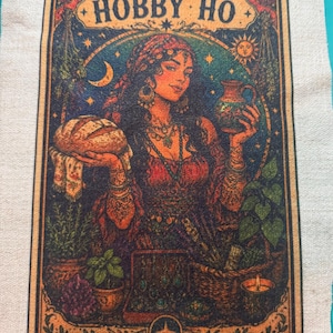 May include: A rectangular fabric panel featuring a printed illustration of a person holding a loaf of bread and a pitcher. The text HOBBY HO is at the top. The scene includes plants, a basket, a candle, and celestial symbols in dark, warm tones.