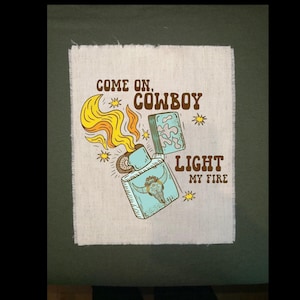 May include: A rectangular patch with a vintage-style illustration. The design features a turquoise lighter with a bull skull, emitting yellow and orange flames. Text reads "Come on, Cowboy, Light My Fire" in brown.