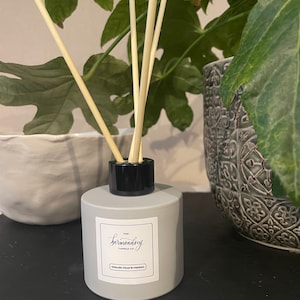 May include: A gray diffuser bottle with a black cap and wooden reeds. The label reads "The Bermondsey Candle Co." and "English Pear & Freesia." The diffuser is placed in front of green leafy plants.
