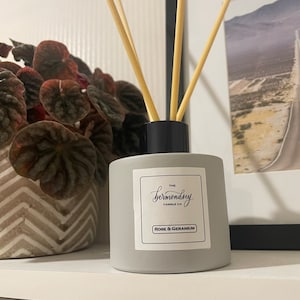 Puede incluir: Una botella difusora gris con tapa negra y palitos de madera. La botella tiene una etiqueta blanca con el texto "THE bermondsey CANDLE CO. ROSE & GERANIUM".