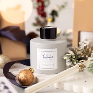 Könnte beinhalten: Eine graue Glasdiffuserflasche mit schwarzem Deckel und weißem Etikett. Das Etikett trägt die Aufschrift "The bermending Candle Co." und "Precious Presents". Die Flasche ist von dekorativen Gegenständen umgeben, darunter ein kleiner künstlicher Weihnachtsbaum, ein goldener Weihnachtsbaumschmuck und weiße Diffusorsticks.
