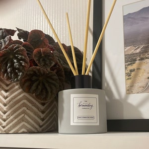 May include: A grey diffuser bottle with a black lid and wooden sticks. The bottle has a white label with the text "The surrounding" on it. The diffuser is sitting on a shelf with a potted plant and books.
