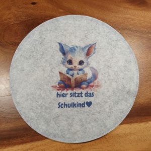 Könnte beinhalten: Ein runder, hellgrauer Filzuntersetzer mit einer Aquarellillustration eines blau-weißen Wesens, das ein Buch liest. Der Text "hier sitzt das Schulkind" ist unter der Illustration aufgedruckt.