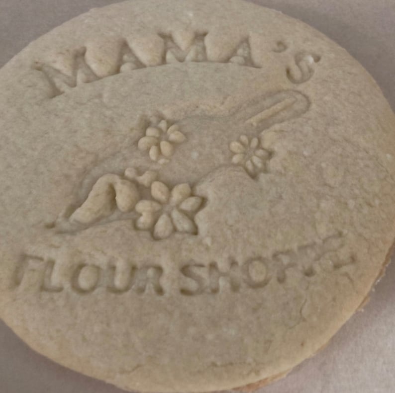 K&ouml;nnte beinhalten: Nahaufnahme eines einfachen, runden, hellbraunen Kekses mit den Worten "Mama's Flour Shoppe" und einem floralen Muster, das in die Oberfl&auml;che gepr&auml;gt ist.