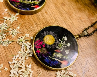 May include: Two gold-toned necklaces with round pendants featuring pressed flowers and greenery encased in clear resin. The pendants are set against a black background.