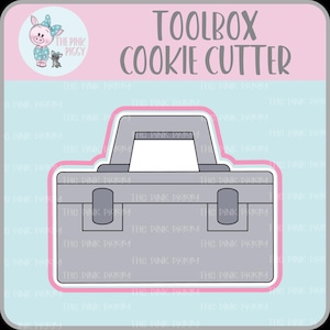 May include: A gray toolbox cookie cutter with a handle and two latches. The text TOOLBOX COOKIE CUTTER is printed at the top. A circular logo featuring a cartoon pig and the text THE PINK PIGGY is visible in the upper left corner.