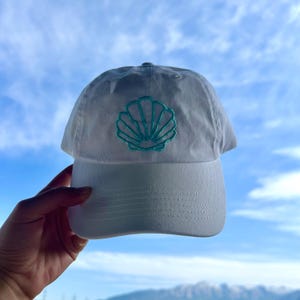 May include: A white baseball cap with a teal seashell embroidered on the front. The cap has a curved brim and a fabric strap closure. The background is a blue sky with clouds.