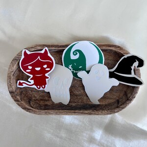 May include: A wooden tray holds several Halloween-themed stickers. Included are a red devil, a green and white swirl, a white skull, a white ghost, and a black witch hat. The stickers are arranged on a wooden tray.