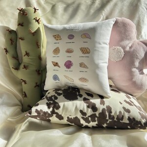 May include: A collection of decorative pillows in various shapes and patterns. Includes a white pillow with seashell illustrations and text, a cowhide print pillow, a pink flower-shaped pillow, and a green cactus-shaped pillow.