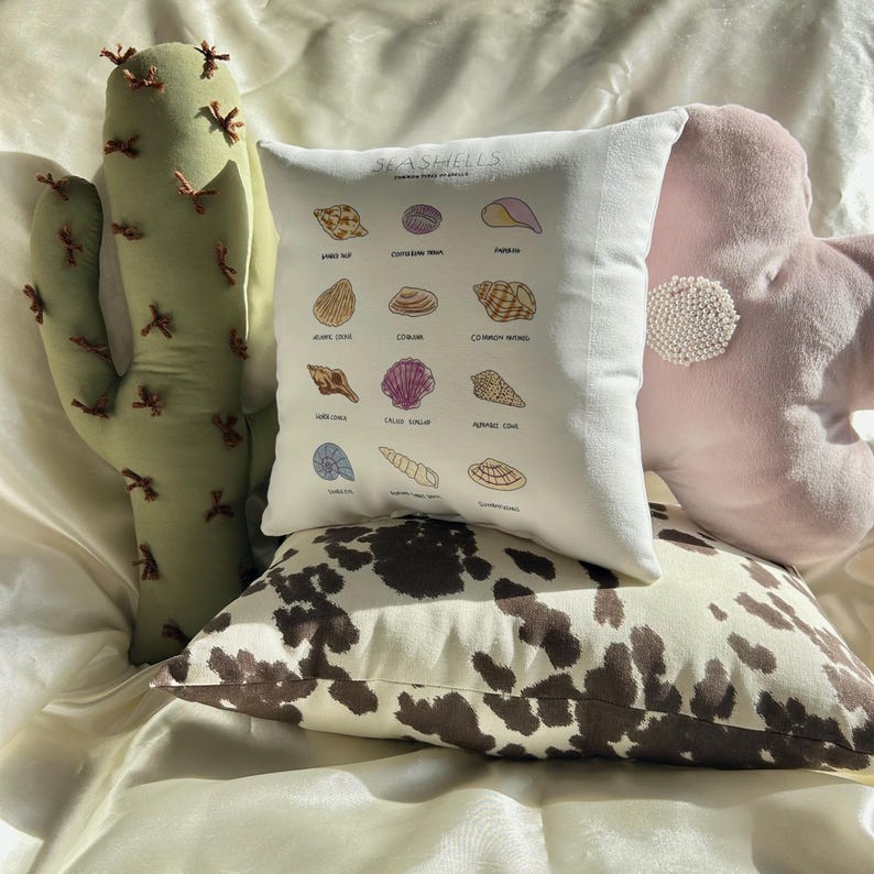 May include: A collection of decorative pillows in various shapes and patterns. Includes a white pillow with seashell illustrations and text, a cowhide print pillow, a pink flower-shaped pillow, and a green cactus-shaped pillow.