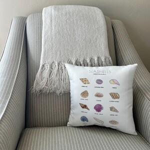 May include: A white decorative pillow with a seashell chart, resting on a striped armchair. A gray fringed throw blanket is draped over the chair. The pillow features various seashell illustrations and the text "SEASHELLS".