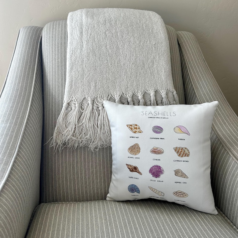 May include: A white decorative pillow with a seashell chart, resting on a striped armchair. A gray fringed throw blanket is draped over the chair. The pillow features various seashell illustrations and the text "SEASHELLS".