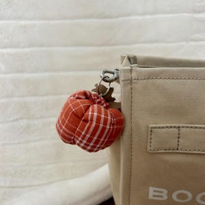 May include: A small, orange and white plaid pumpkin charm hangs from a silver keyring attached to a beige tote bag. The pumpkin charm is made of fabric and has a stem detail. The tote bag has a rectangular shape.