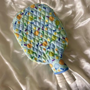Puede incluir: Una funda acolchada para raqueta de pickleball en color verde azulado claro, estampada con varias raquetas de pickleball en amarillo, azul y verde. El mango está envuelto en cinta de agarre blanca y la funda tiene un ribete azul.