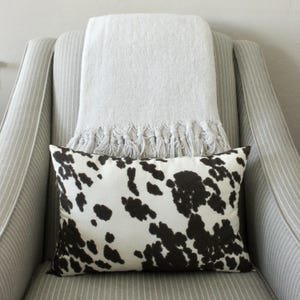 Puede incluir: Un cojín decorativo con estampado de piel de vaca en blanco y negro descansa sobre un sillón a rayas. Una manta gris claro con flecos está colocada sobre el respaldo de la silla, creando un diseño interior acogedor y de tonos neutros.