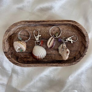 Peut inclure: Quatre porte-clés en forme de coquillages avec des attaches argentées sont présentés sur un plateau ovale en bois. Les coquillages varient en taille, forme et couleur, notamment blanc, marron et beige. Chaque porte-clés a une coquille différente et une petite perle ou pierre décorative.
