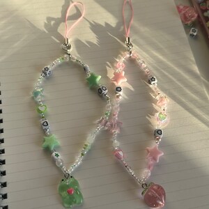 May include: Two phone charms with beaded chains and charms. One charm features a green frog with a pink heart and the other charm features a pink peach. Both charms have white beads, pink and green stars, and hearts.