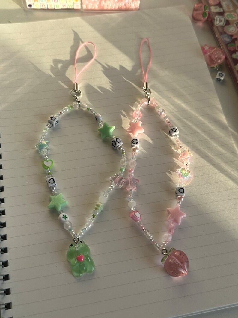 May include: Two phone charms with beaded chains and charms. One charm features a green frog with a pink heart and the other charm features a pink peach. Both charms have white beads, pink and green stars, and hearts.