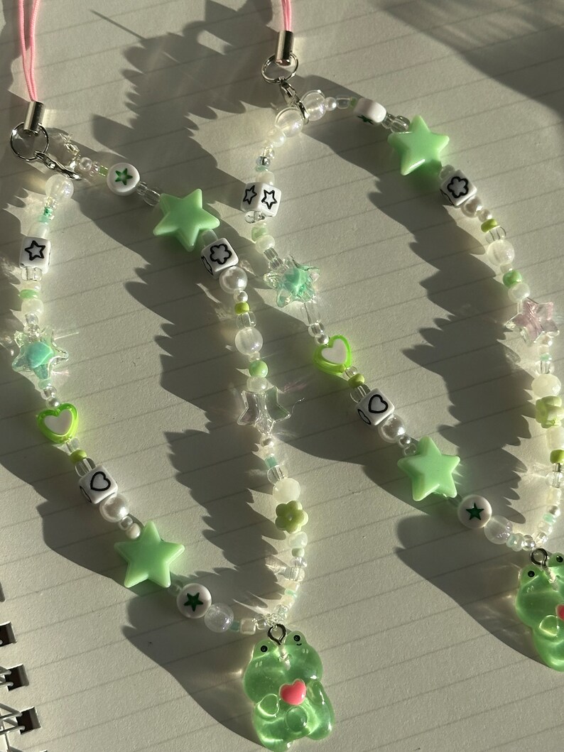 May include: Two green frog charm phone straps with white and green beads, stars, hearts, and flowers.