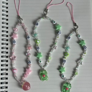 May include: Three phone charms with green and pink beads, stars, hearts, and small charms. The charms are attached to pink strings. The charms feature a strawberry, a frog, and a frog.