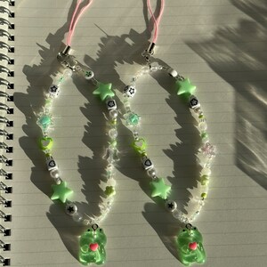 May include: Two green frog charm phone charms with pink string phone straps. The charms are made with green, white, and clear beads, stars, hearts, and flowers.