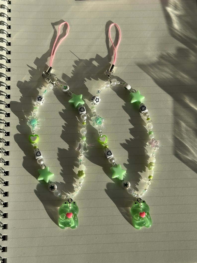 May include: Two green frog charm phone charms with pink string phone straps. The charms are made with green, white, and clear beads, stars, hearts, and flowers.