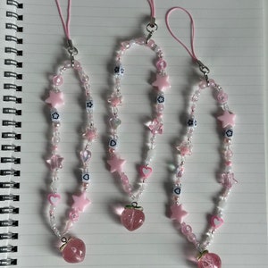 May include: Three pink and white beaded phone charms with star and heart charms and a pink strawberry charm at the bottom.
