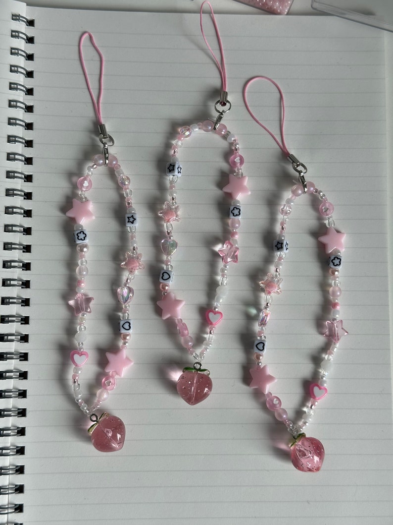May include: Three pink and white beaded phone charms with star and heart charms and a pink strawberry charm at the bottom.