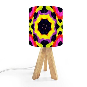 May include: A small table lamp with a colorful, abstract lampshade. The shade features a vibrant pattern of yellow, pink, purple, and black. The lamp has a natural wood tripod base and a white power cord.