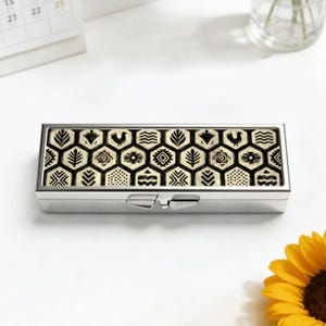 May include: A rectangular silver pill box with a decorative lid featuring a black and cream geometric pattern. The pattern includes stylized birds, leaves, and other shapes within a honeycomb design. The box has a hinged closure.