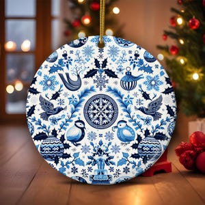 May include: Round ceramic Christmas ornament with a blue and white floral design. The ornament features birds, snowflakes, and other festive elements. It is hanging by a gold string and is approximately 4 inches in diameter.