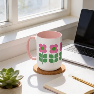 a white mug with pink flowers and green leaves, placed on a cork coaster next to a laptop and a pen on a desk.