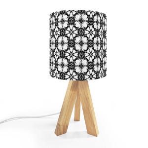 May include: A small table lamp with a cylindrical lampshade featuring a black and white floral pattern. The lampshade sits atop a light-colored, wooden tripod base. A white power cord extends from the base.